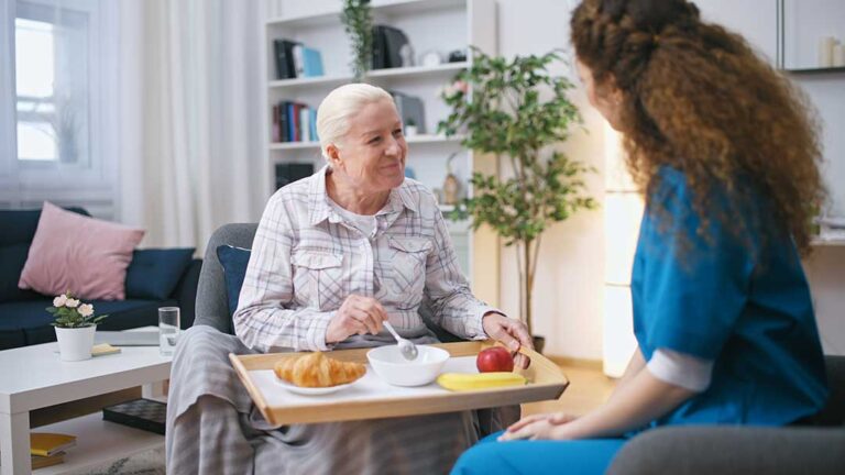Older adult and family member organize labeled clothing, toiletries, photographs, and a small lamp in a bedroom.